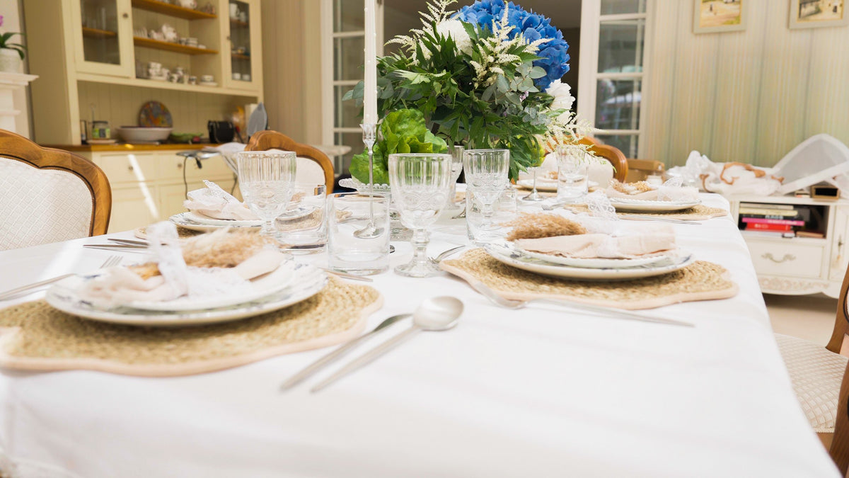A dining table set for four with woven rattan placemats, blue and white patterned plates, lace-tied napkins, embossed glassware and a blue hydrangea centrepiece