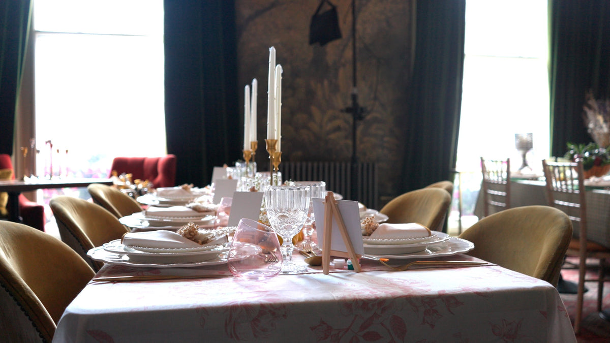 An elegant dining room with a long styled table, white taper candles in gold candlesticks and velvet chairs beside tall windows