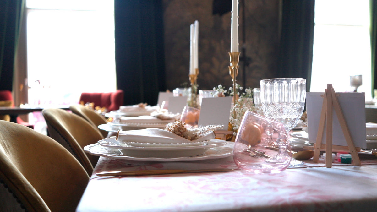 A long dining table dressed in a pink toile tablecloth with scalloped white plates, gold cutlery, taper candles and mustard velvet chairs
