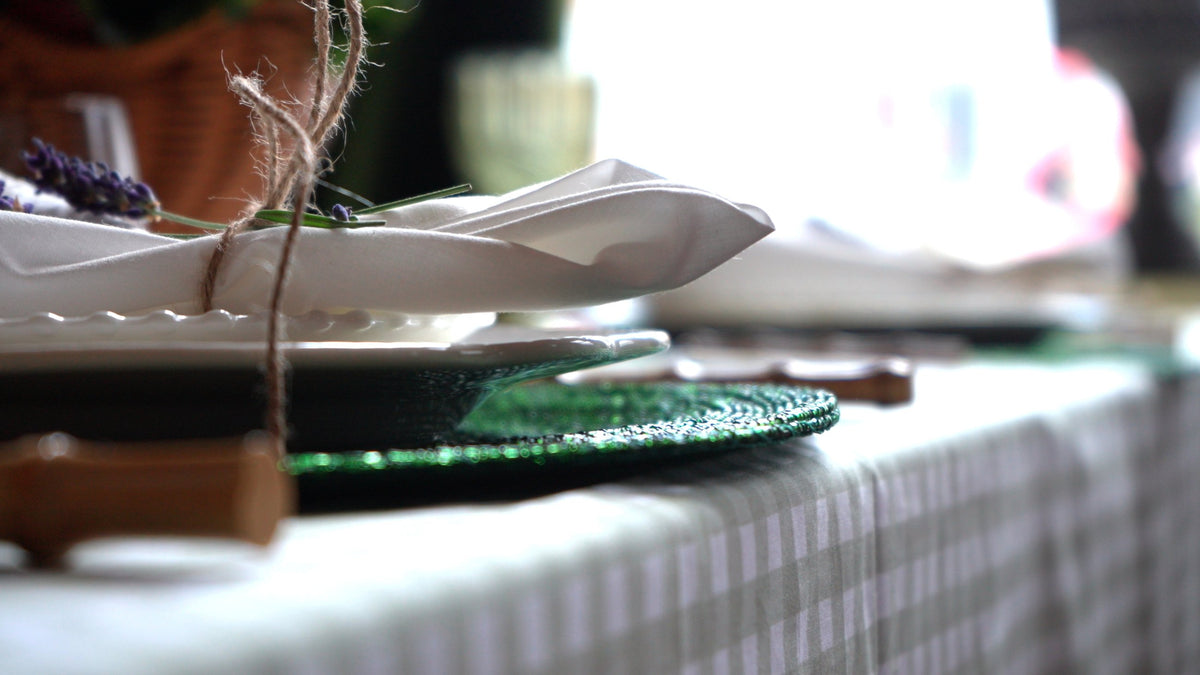 A close-up of a place setting with a folded white napkin tied in twine with sprigs of lavender, on a green beaded charger and gingham tablecloth